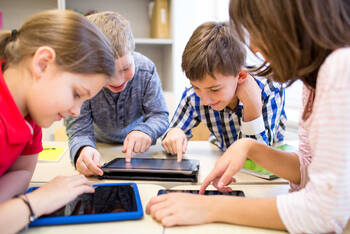 Drie kinderen zitten met tablets rond een tafel. Een volwassen zit er ook bij.