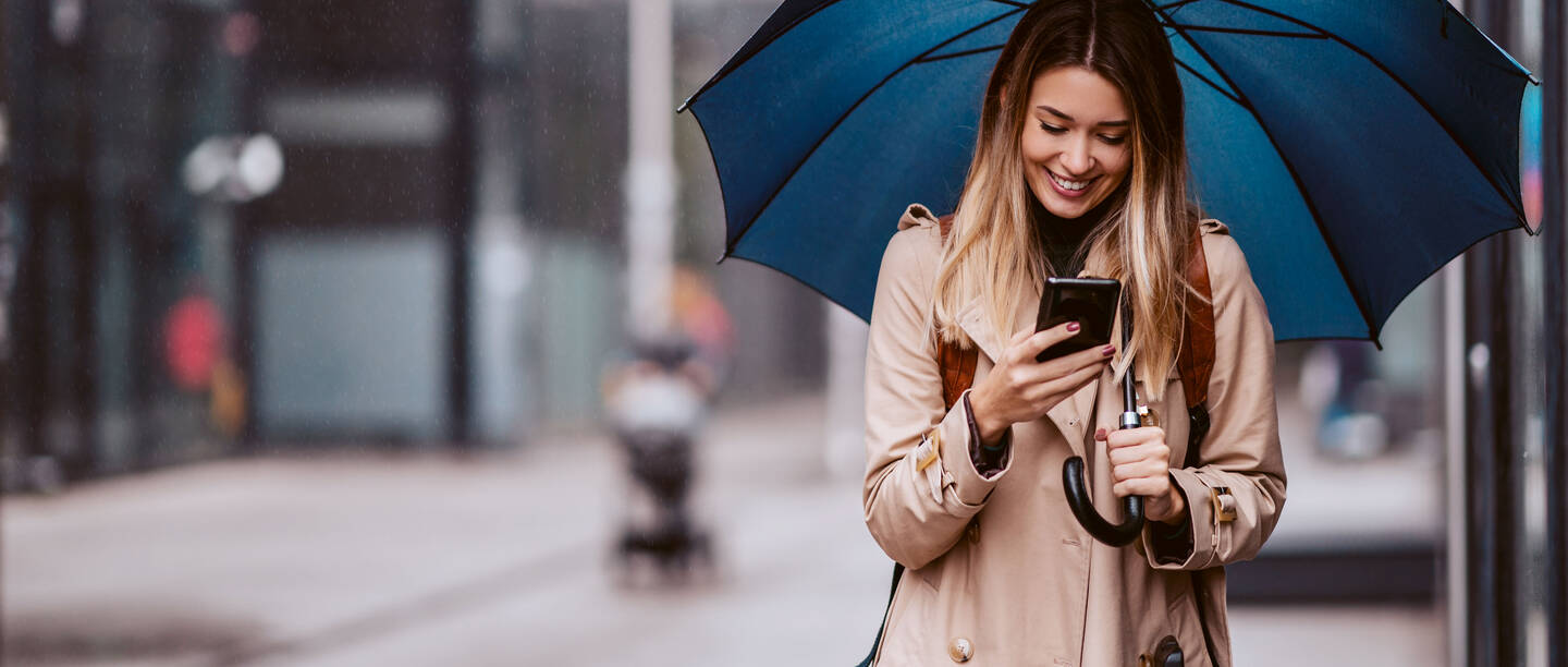 Een jonge vrouw in een beige trenchcoat met paraplu kijkt glimlachend naar haar smartphone