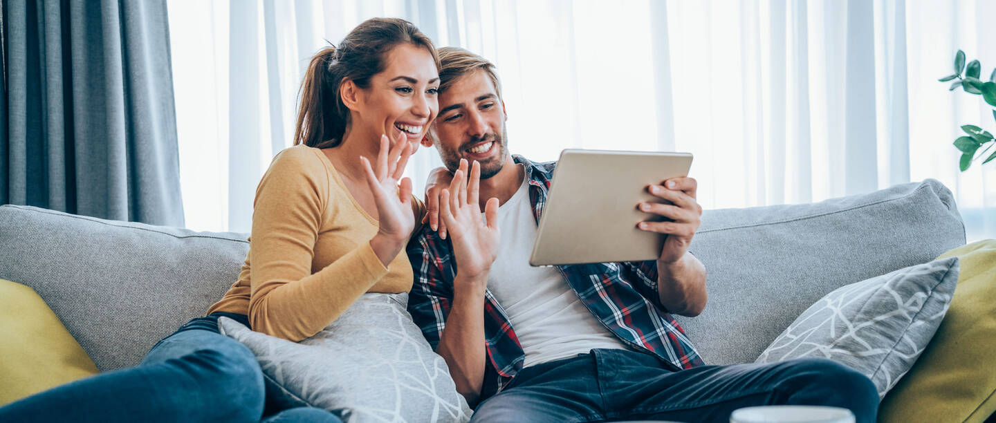 Een jong stel zit op de bank. Ze houden de tablet in hun handen en zwaaien ernaar.