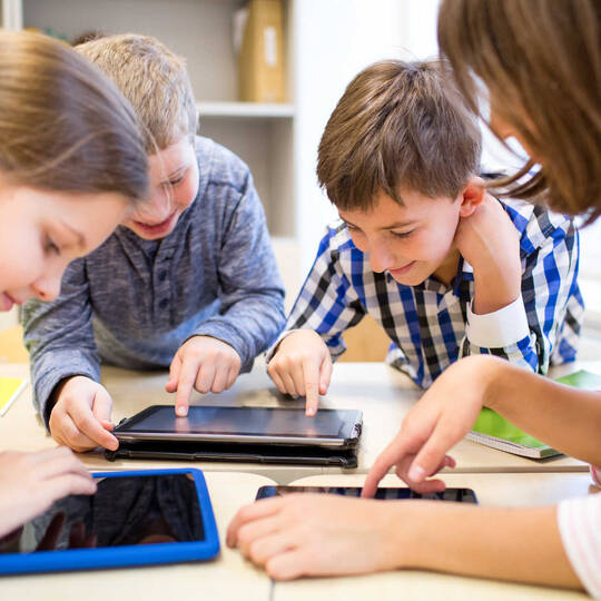 Tablets in het onderwijs. Drie kinderen zitten met tablets rond een tafel. Een volwassen zit er ook bij.